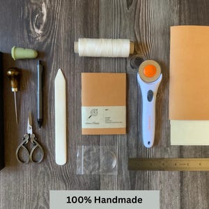 A travelers notebook insert surrounded by all of the tools and materials used to make the handmade notebook inserts.  Some of the tools include the vegan leather, animal-free parchment paper, linen thread, curved needle, bone folder and scissors.