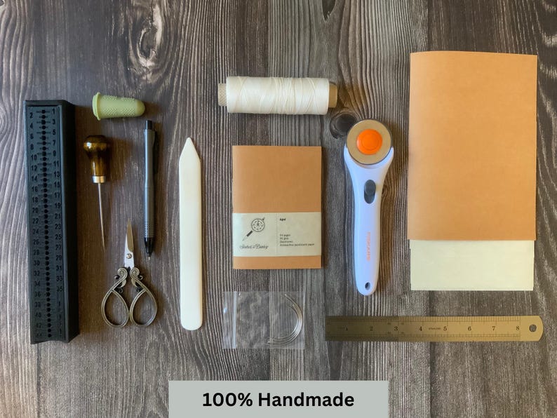 A travelers notebook insert surrounded by all of the tools and materials used to make the handmade notebook inserts.  Some of the tools include the vegan leather, animal-free parchment paper, linen thread, curved needle, bone folder and scissors.