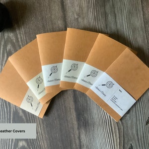 A set of 6 travelers notebook inserts fanned out over a wood surface.  The paper labels on each notebook insert match the page colors inside.  The covers are made of a brown vegan leather, also known as washable paper.