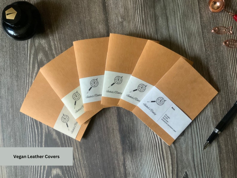 A set of 6 travelers notebook inserts fanned out over a wood surface.  The paper labels on each notebook insert match the page colors inside.  The covers are made of a brown vegan leather, also known as washable paper.