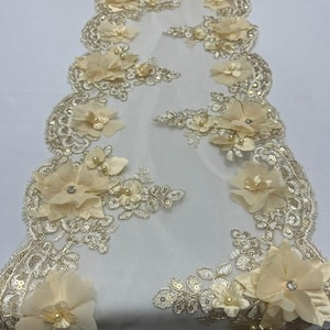 May include: Ivory lace table runner with floral and sequin embellishments. The runner features large, three-dimensional fabric flowers with pearl and rhinestone accents, and intricate lace detailing along the edges. The design is symmetrical and suitable for weddings or special events.