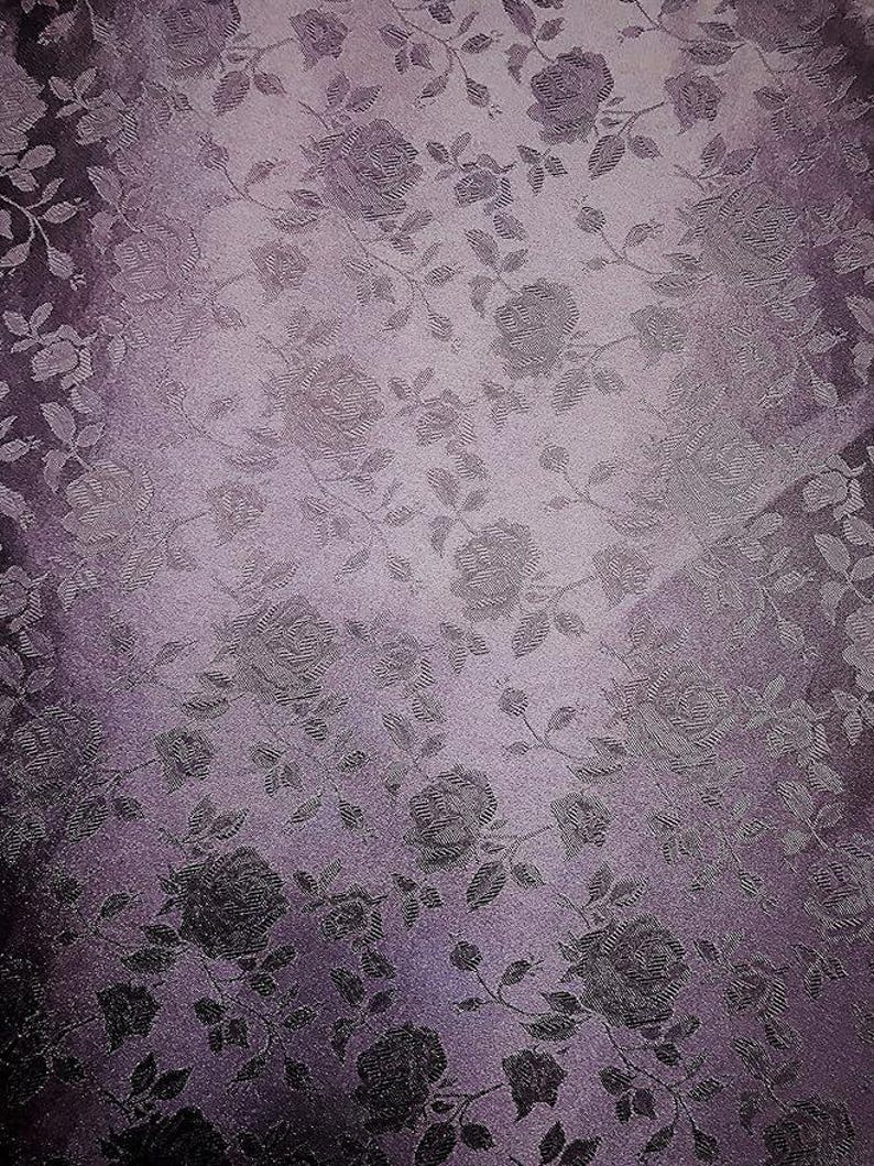 May include: A textured fabric with a floral pattern featuring roses and leaves in shades of purple and gray. The design has a subtle sheen, creating a vintage or romantic aesthetic. The fabric's color gradient adds depth to the overall design.
