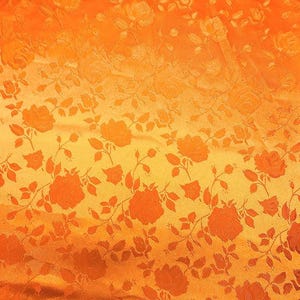 May include: Close-up of orange fabric with a floral pattern. The design features repeating rose motifs with stems and leaves. The fabric has a textured appearance and a gradient effect, transitioning from a darker orange at the top to a lighter yellow-orange at the bottom.