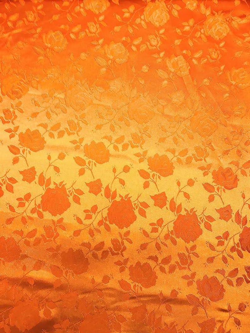 May include: Close-up of orange fabric with a floral pattern. The design features repeating rose motifs with stems and leaves. The fabric has a textured appearance and a gradient effect, transitioning from a darker orange at the top to a lighter yellow-orange at the bottom.