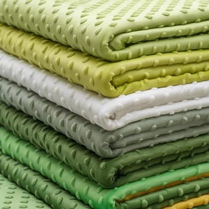 May include: A stack of plush blankets in various shades of green and white. Each blanket has a textured, raised dot pattern. The colors range from light lime to deep forest green, creating a visually appealing display of soft textiles.