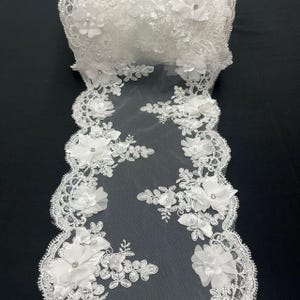 May include: White lace fabric with floral embellishments. The fabric features delicate, three-dimensional flowers, small beads, and a scalloped edge. The lace is rolled up, showcasing the intricate details and texture of the design. Ideal for bridal or decorative projects.
