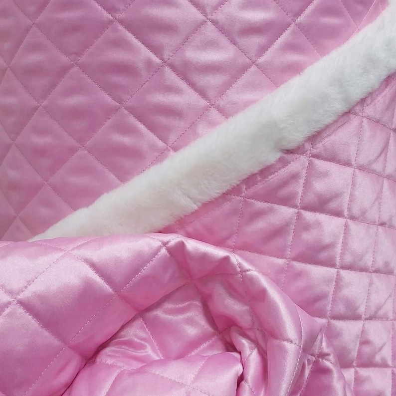 May include: Close-up of a pink quilted fabric with a diamond pattern. A white, fluffy trim runs diagonally across the image. The fabric has a satin sheen, suggesting a soft and luxurious texture. The overall aesthetic is delicate and feminine.