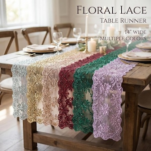 May include: A collection of floral lace table runners, 14 inches wide, in silver, gold, champagne, burgundy, emerald green, and lavender. The runners feature intricate sequined floral patterns with scalloped edges, displayed on a wooden table.