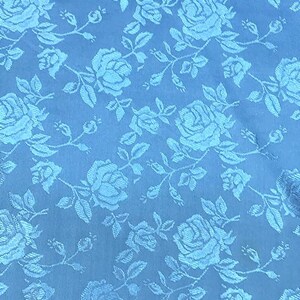May include: A close-up of a light blue fabric with a repeating pattern of roses and leaves. The roses are in full bloom, and the leaves are detailed. The fabric has a subtle sheen, giving it a luxurious appearance.