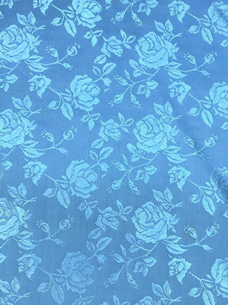 May include: A close-up of a light blue fabric with a repeating pattern of roses and leaves. The roses are in full bloom, and the leaves are detailed. The fabric has a subtle sheen, giving it a luxurious appearance.