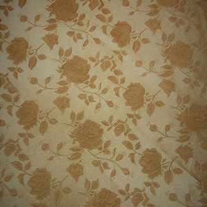 May include: Close-up of a fabric with a repeating pattern of roses and leaves. The roses and foliage are a warm, golden-brown color against a cream-colored background. The fabric has a subtle sheen.