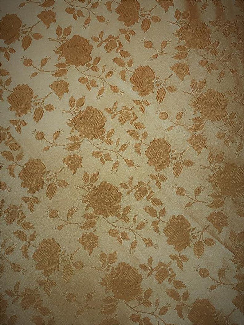 May include: Close-up of a fabric with a repeating pattern of roses and leaves. The roses and foliage are a warm, golden-brown color against a cream-colored background. The fabric has a subtle sheen.