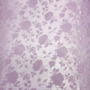 May include: A close-up of a lavender fabric with a repeating rose pattern. The roses and stems are a darker shade of lavender, creating a subtle contrast against the lighter background. The fabric has a slight sheen.
