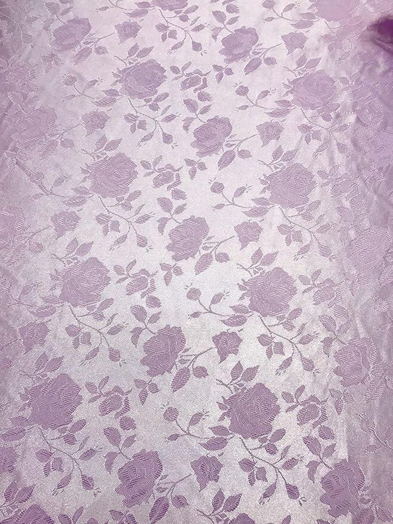 May include: A close-up of a lavender fabric with a repeating rose pattern. The roses and stems are a darker shade of lavender, creating a subtle contrast against the lighter background. The fabric has a slight sheen.