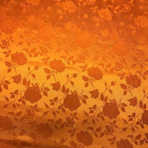 May include: Close-up of a fabric with a repeating rose pattern. The fabric has a warm orange color with darker orange rose and leaf details. The texture appears to be slightly raised, creating a subtle pattern.