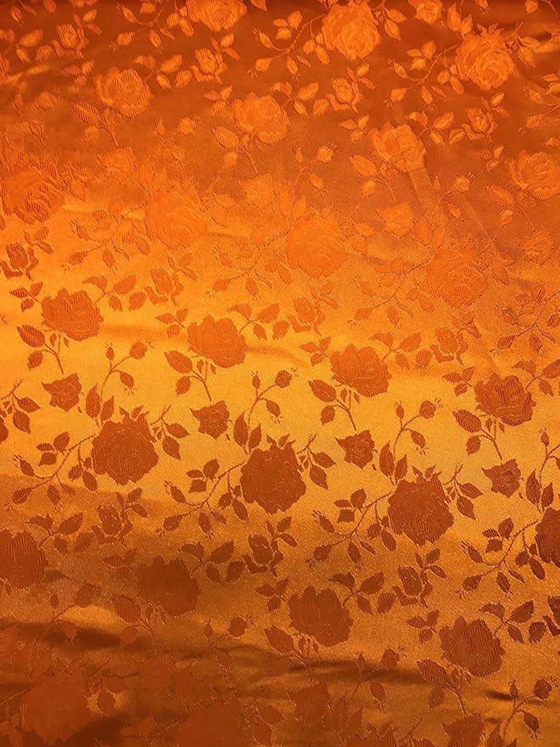 May include: Close-up of a fabric with a repeating rose pattern. The fabric has a warm orange color with darker orange rose and leaf details. The texture appears to be slightly raised, creating a subtle pattern.