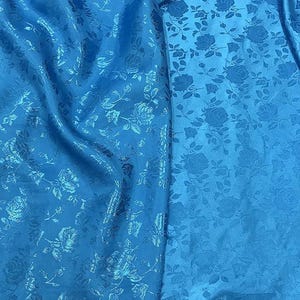 May include: Bright blue fabric with a floral rose pattern. The fabric has a slight sheen and is draped, showing the texture and design. The roses are subtly embossed, adding depth to the textile.