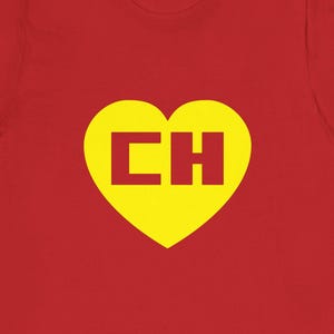 May include: A red short-sleeved t-shirt featuring a large yellow heart graphic centered on the chest. Inside the yellow heart, the letters CH are printed in a bold, dark red block font. The shirt has a crew neckline and a standard straight hem.