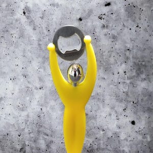 May include: A yellow bottle opener shaped like a person holding a silver bottle opener above its head.