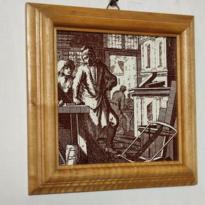 May include: A framed tile featuring a black and white illustration of a workshop scene. The illustration shows a man and a woman working at a table with tools and other objects around them. The tile is framed in a light brown wooden frame.