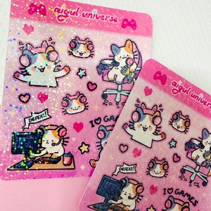 Puede incluir: Dos hojas rosas de pegatinas con lindos gatos de dibujos animados que llevan auriculares y juegan a videojuegos. Las pegatinas tienen un acabado hologr&aacute;fico e incluyen el texto "aigul universe" y "Me encantan los juegos".