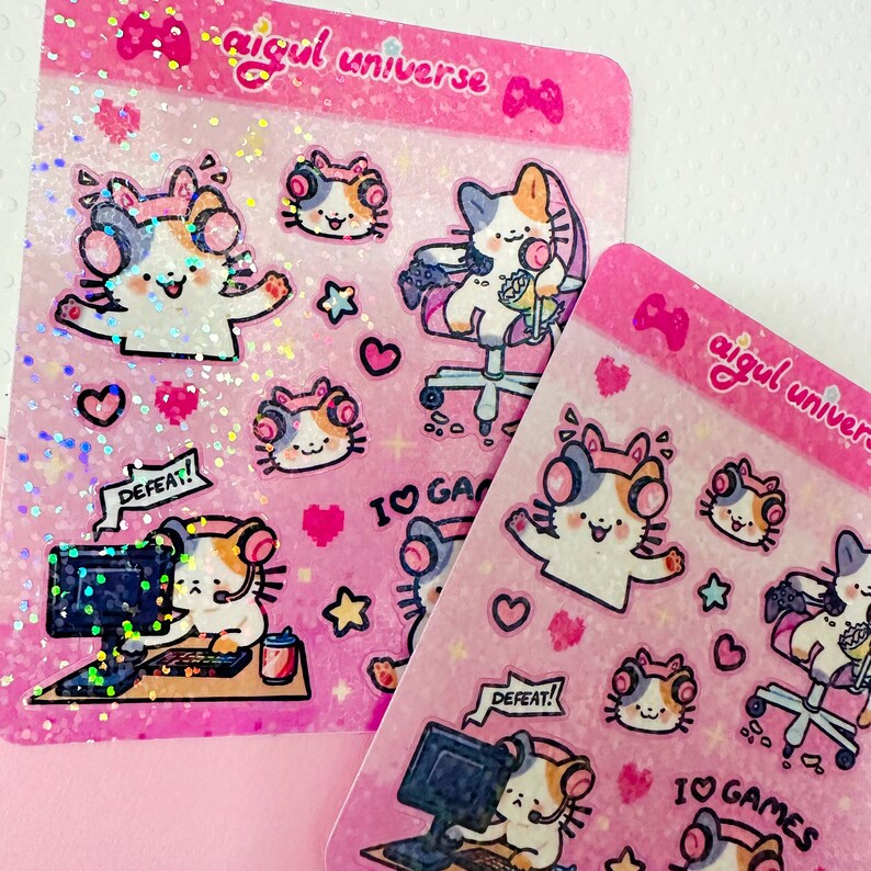 Puede incluir: Dos hojas rosas de pegatinas con lindos gatos de dibujos animados que llevan auriculares y juegan a videojuegos. Las pegatinas tienen un acabado hologr&aacute;fico e incluyen el texto "aigul universe" y "Me encantan los juegos".