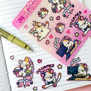 Puede incluir: Un conjunto de pegatinas hologr&aacute;ficas con lindos gatos de dibujos animados que usan auriculares y juegan videojuegos. Las pegatinas tienen un fondo rosa con brillo y el texto "I♡GAMES" y "DEFEAT!"