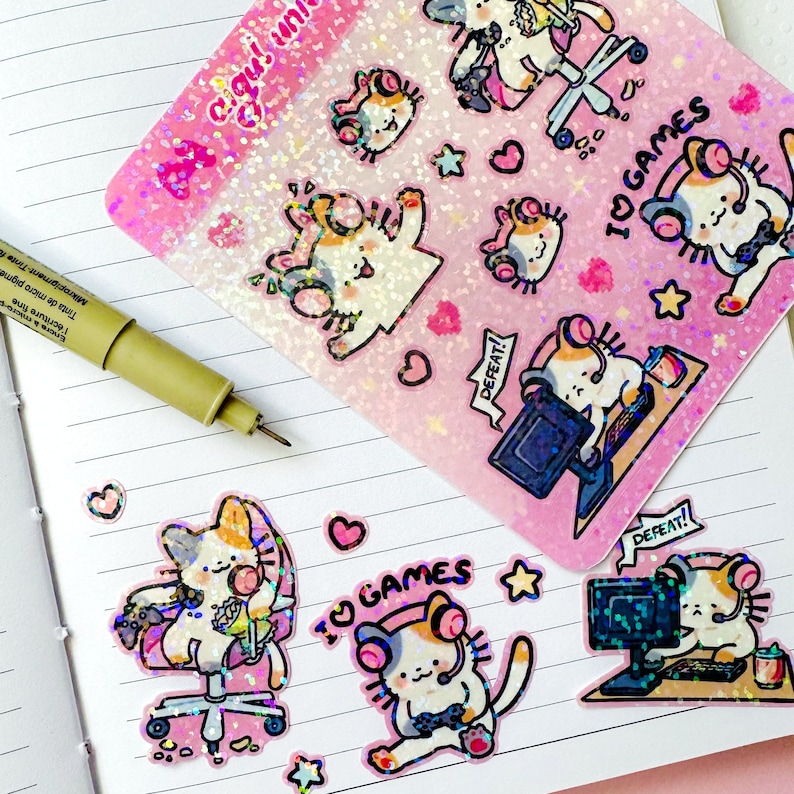 Puede incluir: Un conjunto de pegatinas hologr&aacute;ficas con lindos gatos de dibujos animados que usan auriculares y juegan videojuegos. Las pegatinas tienen un fondo rosa con brillo y el texto "I♡GAMES" y "DEFEAT!"