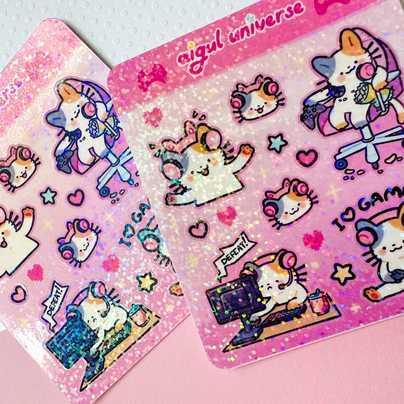Puede incluir: Dos hojas de pegatinas hologr&aacute;ficas rosas con lindos gatos de dibujos animados que llevan auriculares y sostienen mandos de juego. Las pegatinas tienen un acabado brillante e incluyen frases como "DEFEAT!" e "I &hearts; GAMING".