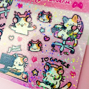 Puede incluir: Una hoja de pegatinas hologr&aacute;ficas rosas con lindos gatos de dibujos animados con auriculares y accesorios de juego. Las pegatinas dicen "Me encantan los juegos" y "&iexcl;Derrota!"