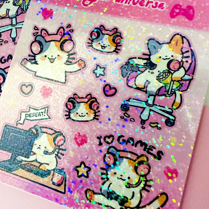 Puede incluir: Una hoja de pegatinas hologr&aacute;ficas rosas con lindos gatos de dibujos animados con auriculares y accesorios de juego. Las pegatinas dicen "Me encantan los juegos" y "&iexcl;Derrota!"