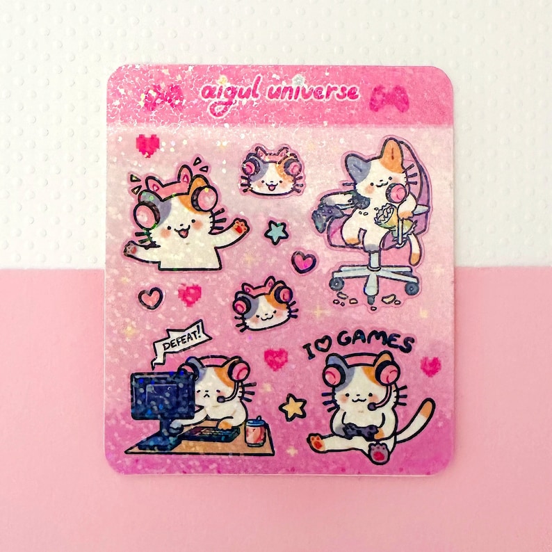 Puede incluir: Una hoja de pegatinas rosa y blanca con un fondo brillante que presenta una variedad de gatos de dibujos animados lindos jugando videojuegos. La hoja incluye el texto "aigul universe" y "Me encantan los juegos".