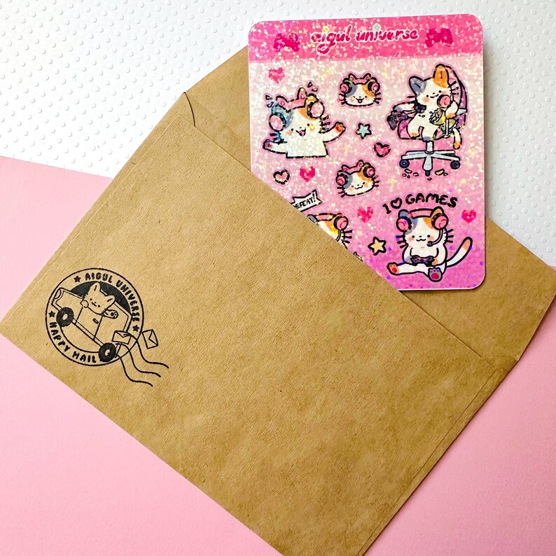 Puede incluir: Un sobre rosa y blanco con una hoja de pegatinas que presenta un gato de dibujos animados con auriculares y el texto "Aigul Universe" y "Me encantan los juegos".