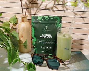 Può includere: Una bustina verde con il testo "Gut Teas™ Emerald Super Latte Matcha - Ashwagandha - Maca 30 servings | 1 daily for each bag". Un bicchiere di latte matcha verde con ghiaccio e una cannuccia di vetro. Occhiali da sole con montatura marrone e lenti verdi.