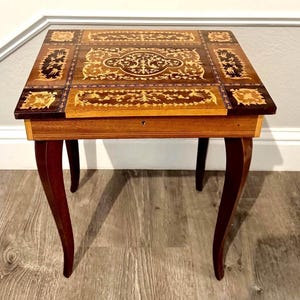 May include: A square wooden side table featuring intricate marquetry inlay on the top surface. The table has four curved legs and a light brown wood frame with a small keyhole centered on the front apron beneath the tabletop.