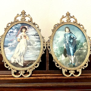 May include: Two oval brass frames with ornate scrollwork detailing, each containing a portrait. The left frame holds a portrait of a person in a white dress with a red ribbon. The right frame holds a portrait of a person in a blue suit.