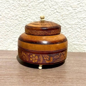 May include: A round wooden lidded box with a polished brown finish. The lid features a small cream-colored knob and a geometric Greek key pattern. The base displays an intricate floral design etched into the wood, supported by small cream feet.