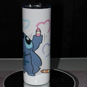 Pu&ograve; includere: Un bicchiere in acciaio inossidabile bianco con un personaggio dei cartoni animati blu che disegna cuori con un pastello rosa. Il personaggio &egrave; del film Disney Lilo & Stitch.