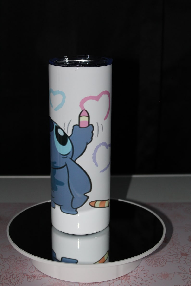 Pu&ograve; includere: Un bicchiere in acciaio inossidabile bianco con un personaggio dei cartoni animati blu che disegna cuori con un pastello rosa. Il personaggio &egrave; del film Disney Lilo & Stitch.