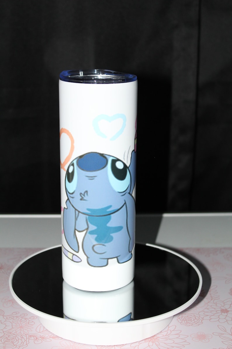 Pu&ograve; includere: Un bicchiere in acciaio inossidabile bianco con un personaggio Stitch blu di Lilo & Stitch. Il personaggio &egrave; disegnato in stile cartone animato e tiene una matita rosa. Lo sfondo &egrave; bianco con cuori e cerchi colorati.