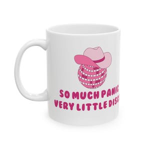 May include: White ceramic mug featuring a pink disco ball wearing a pink cowboy hat. The mug has the text "So much panic, very little disco" printed on it.