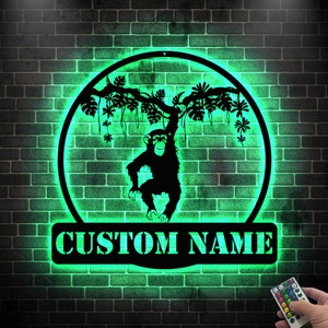 Może przedstawiać: Zielony, podświetlany znak metalowy z sylwetką małpy wiszącej na winorośli. Znak ma miejsce na spersonalizowane imię, które jest pokazane jako "CUSTOM NAME".