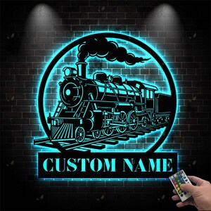 Peut inclure: Silhouette noire d'un train avec une lueur néon bleue, encadrée dans un cercle. Le design comprend le texte "CUSTOM NAME" sur une base rectangulaire. L'image est fixée sur un mur de briques.