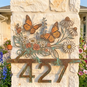 May include: A metal house number plaque displaying the digits 427 in a rustic bronze finish with a verdigris patina. The design features two orange monarch butterflies, blue floral sprigs, and white daisy-like flowers with green leaves.