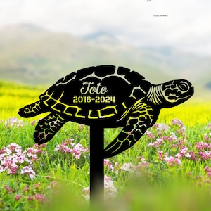 Op de afbeelding: Zwarte metalen silhouet van een zeeschildpad met de naam "Toto" en de data "2016-2024" gegraveerd op de schild. De schildpad is gemonteerd op een zwarte metalen paal, geschikt voor buitengebruik.