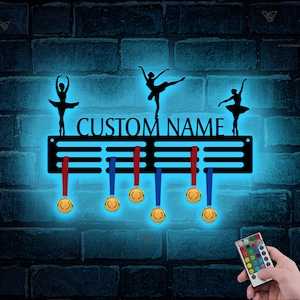 May include: Black metal medal display rack with a custom name, "CUSTOM NAME", and three ballerina silhouettes. The rack has six hooks for hanging medals and a remote control for changing the color of the LED lights.