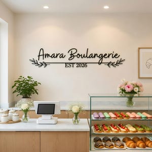 May include: A black metal sign that reads "Amara Boulangerie EST 2026" with leaf accents. The sign is mounted on a white wall above a bakery counter with pastries and flowers.