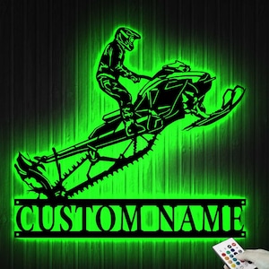 May include: A black metal silhouette of a person riding a snowmobile with the text "CUSTOM NAME" below. The silhouette is lit up in green.