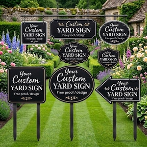 May include: A collection of black custom yard signs in various shapes, including square, oval, and arrow. Each sign features white text that reads "Your Custom YARD SIGN" with "Free proof / design" below. The signs are displayed in a garden setting.