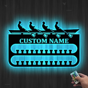 Puede incluir: Una decoración de pared de metal negro con una silueta de un equipo de remo en un bote. La decoración tiene un brillo azul y un espacio para un nombre personalizado. El texto "CUSTOM NAME" es visible en la decoración.